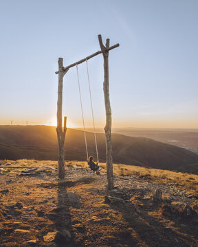 View of a solitary figure on a rustic wooden swing silhouetted against a radiant sunset atop a mountain ridge, casting long shadows, Lous&atilde;, Coimbra, Portugal.