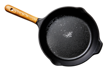 Seasoned black cast iron skillet with a faint sheen, honey oak wooden handle sleeve and a few coarse salt crystals inside isolated on transparent background.
