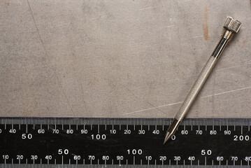 Metal scribe tool on scratched metal surface with steel ruler on industrial metal background.