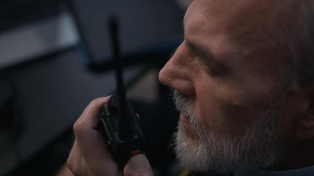 Close view of adult Caucasian male security officer speaking into two way radio while sitting at desk in front of surveillance screens in dark control room