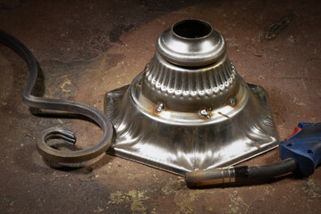 Metal welded lamp base on background with forged hook and welding torch resting on workshop floor background.