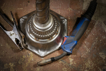 Metal welded lamp base on background with forged hook and welding torch resting on workshop floor background.