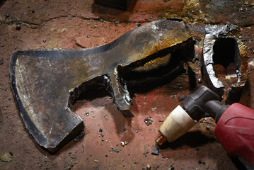 Forged steel axe head blank with black layout markings on wooden anvil block beside welding helmet and plasma cutter torch in blacksmith workshop close up.