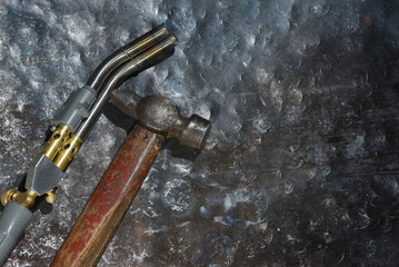 Gas metal heating torch with dual curved nozzles and worn blacksmith hammer placed on hammered steel workshop surface close up.