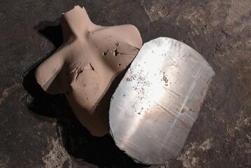 Forged steel armor bra with two hammered domed cups formed on clay mold during blacksmithing process on workshop floor background.