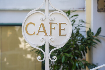View of a white ornate sign announcing 'CAFE' in gold lettering, juxtaposed against a blur of foliage and building facade, Saint Gilgen, Salzburg, Austria.