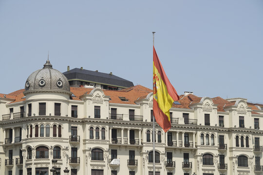 View of the Spanish flag boldly waves against the backdrop of ornate, creamy buildings with terracotta roofs under a clear sky, Santander, Cantabria, Spain.