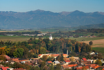 View of a quaint village nestled among vibrant autumn foliage, with a church steeple piercing the skyline against a backdrop of distant, majestic mountains, Necpaly, Zilina Region, Slovakia.