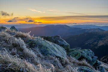 View of the sun setting behind distant mountain peaks, casting a warm glow on the frosted grass and rocks, Location data omitted.