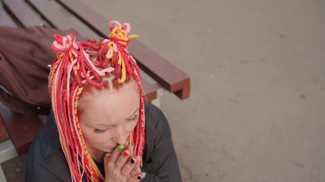 white woman with pink dreadlocks wiping tears with hands, hunched over on wooden bench beside backpack, pavement and park trees in background, intimate portrait of acute sadness and release