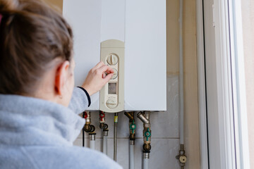 Rear view of a woman in a grey fleece operating a domestic gas boiler, showing everyday home heating adjustment, utility use and energy efficiency in a residential interior. © Alina Mosinyan