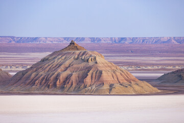 View of a layered, sun-kissed mesa rising dramatically from the stark white earth under a serene sky, its muted hues painting a surreal landscape, Karynzharyk, Mangystau Region, Kazakhstan.