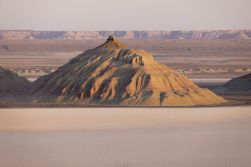View of a solitary mountain rises from the cracked earth, bathed in the warm hues of the setting sun, casting long shadows across the arid landscape, Karynzharyk, Mangystau Region, Kazakhstan.