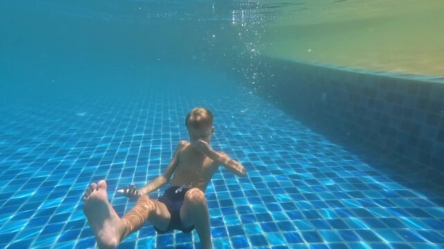 Underwater shot of a child in glasses swimming over a blue mosaic of the bottom with bubbles and sun glare; horizontal video.