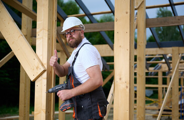 Construction worker using an electric screwdriver while building a wooden frame structure. Man in...