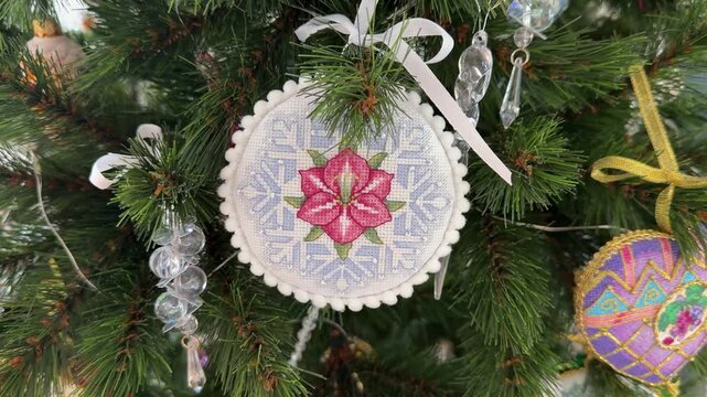A close-up of handmade cross-stitch Christmas ornament featuring a pink flower and blue snowflakes hanging on a Christmas tree. All Christmas ornaments embroidered and made by myself.