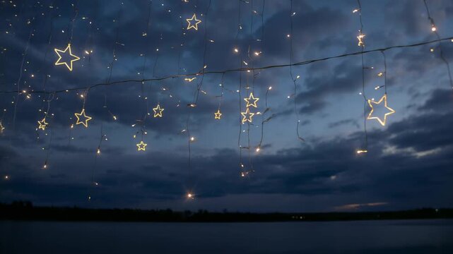 Glowing string of star-shaped lights hanging on wire, swaying and twinkling over calm lake at dusk