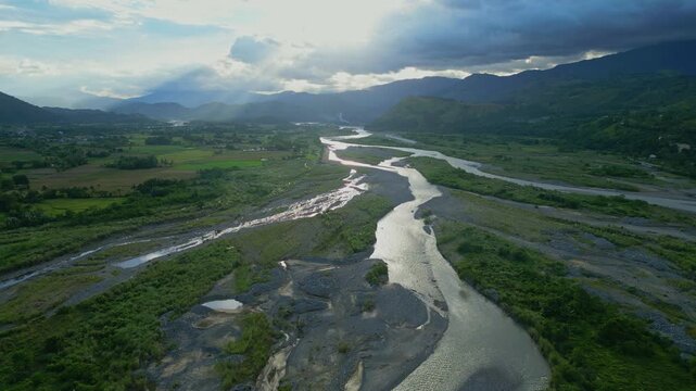 A rising forward aerial of the diverging Magat River glows under sunbeams peeking through cloud breaks Bontoc, Nueva Vizcaya, Philippines