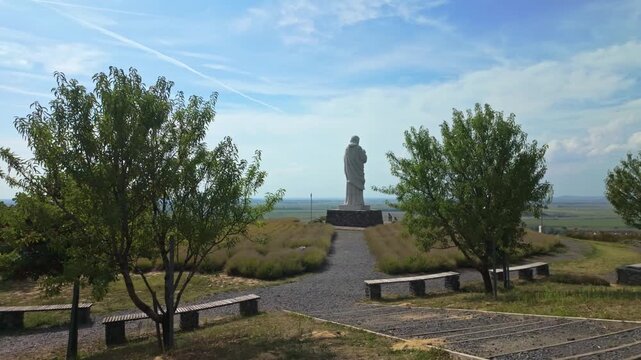 Sideways tracking reveals the &Aacute;ld&oacute; Krisztus statue in Tarcal, Hungary, surrounded by lush greenery, open fields, and a peaceful park with pathways, benches, and a serene European landscape.