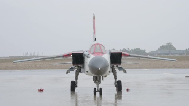 Front view of a military jet aircraft standing on a wet runway during rainy weather with landing gear deployed and airbase background