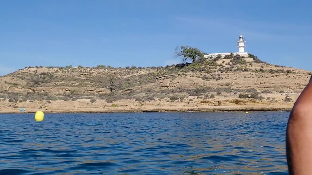 Point of view from a kayak in San Juan bay showing the Cabo de las Huertas lighthouse on a hill with a tree on a sunny summer day in Alicante Spain