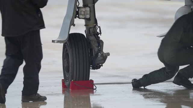 Ground crew member crouching beside aircraft landing gear placing wheel chock on wet runway during ground operation