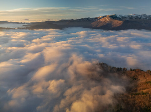 Aerial view of a sea of clouds blankets the landscape below Jelenska skala, with distant snow-capped mountains catching the golden light, Jelenska skala, Banska Bystrica Region, Slovakia.