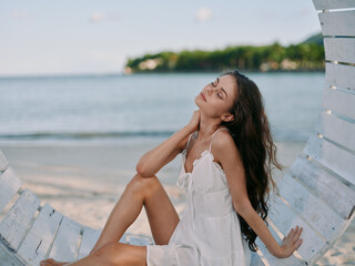 A serene young woman sits in a white sundress on a wooden swing by the shore, sunlit breeze, calm sea, and tranquil coastal beauty.