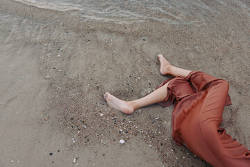 A lone person lies on wet sand near the shoreline, legs extended in rust-colored pants, toes...