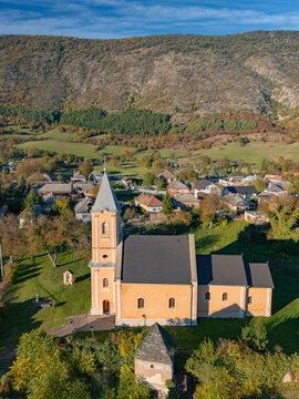 Aerial view of the Church of St. John the Baptist basking in golden sunlight amidst the village and the backdrop of a rugged mountain, Hrhov, Kosice Region, Slovakia.