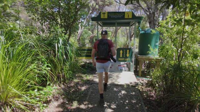 A hiker cleans his boots at a cleaning station to prevent the spread of Kauri dieback disease in a forest in New Zealand. The man is wearing a backpack and carrying a camera.