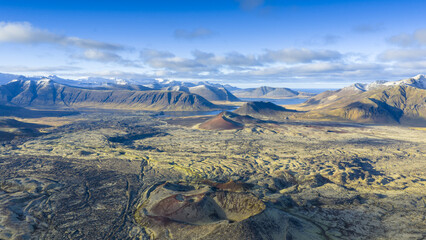 Aerial view of stark volcanic cones punctuate the rugged landscape under a vast sky, where snow-dusted peaks meet the distant ocean, iCELAND, Iceland.