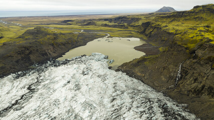 Aerial view of glacial ice meeting a silt-laden lake, contrasted by dark cliffs and distant plains under an overcast sky, Solheimajokull, Myrdalshreppur, Iceland.