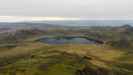 Aerial view of a serene lake cradled by rugged, moss-kissed hills under a vast, muted sky, the landscape a tapestry of earthy tones meeting the distant sea, Solheimajokull, Myrdalshreppur, Iceland.