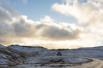 View of a small triangular house nestled in a snowy landscape beneath a dramatic sky of sun-kissed clouds, Snaefellsjokull, Snaefellsbaer, Iceland.