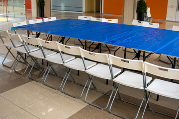 A bright interior scene shows a neat stack of white folding chairs beside a tall black planter holding a lush green topiary, set against a neutral marble-tiled wall in a modern lobby or event space.