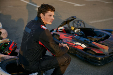 A focused young man in a racing suit sits beside his go-kart, contemplating the upcoming race
