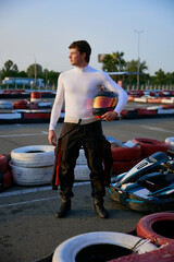 A go-kart racer stands confidently at the karting circuit wearing racing gear and holding a helmet