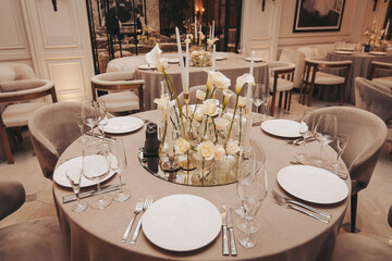 A dining table is set with white plates and glasses. Flowers are in a vase at the center. The room has soft lighting and a stylish arrangement. Chairs are around the table for guests