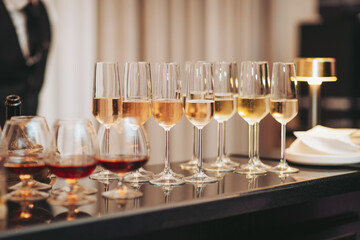 A bartender prepares various drinks for guests. Glasses with different beverages sit on a bar. Guests are expected to arrive soon. The setting is inside a modern venue