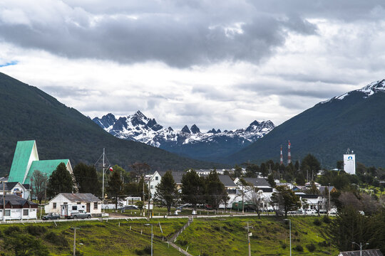 Aerial view of a town nestled in a valley, with snow-capped Dientes de Navarino mountains rising in the background under a cloudy sky, Puerto Williams, Chile.