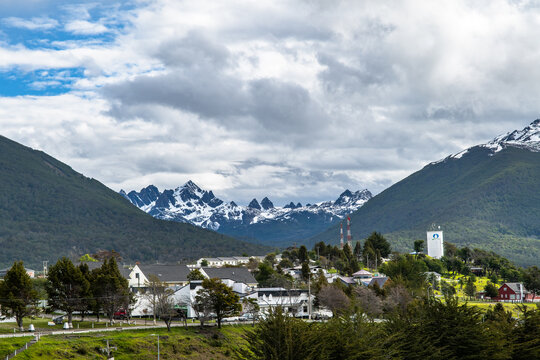 Aerial view of a quaint town nestled amidst verdant hills, with snow-capped Dientes de Navarino mountains piercing the skyline, a serene blend of nature and civilization., Puerto Williams, Chile.