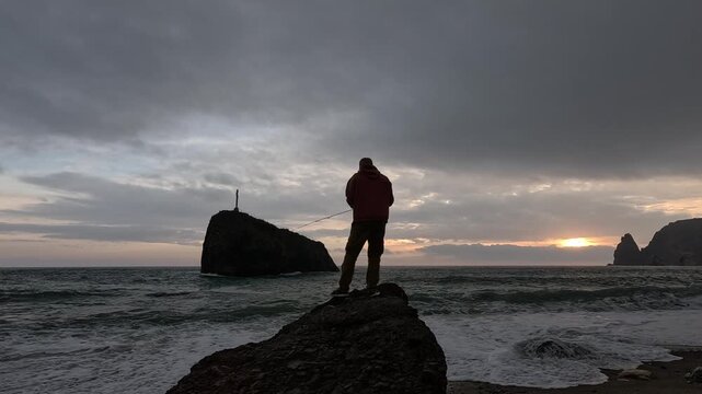 Man fishing ocean, standing on a rock at sunset with dramatic cloudy sky, casting line into rough sea