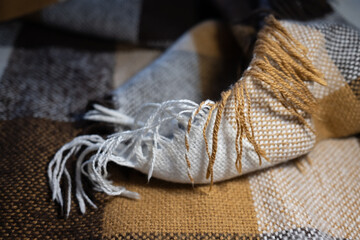 A soft, folded plaid blanket with a checkered pattern in rustic tones. Close-up on the cozy texture and decorative white and beige fringe, creating a warm, hygge atmosphere.