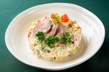 A close-up shot of a gourmet Olivier salad served on a white plate, topped with delicate slices of meat, red caviar, and fresh herbs, against a dark green background