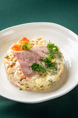 A close-up shot of a gourmet Olivier salad served on a white plate, topped with delicate slices of meat, red caviar, and fresh herbs, against a dark green background