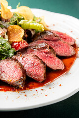 A vibrant, gourmet steak salad is served on a white plate, showcasing perfectly seared, sliced rare beef and a fresh mix of greens, cherry tomatoes, and crispy potato chips