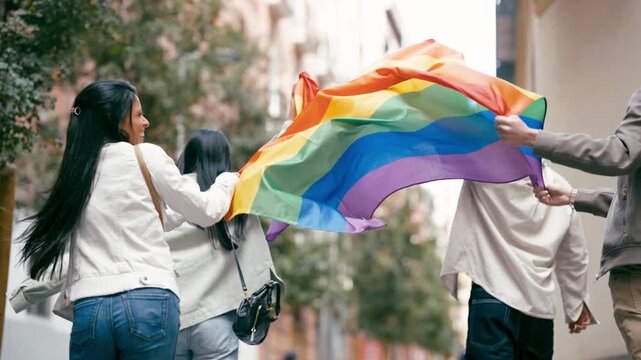 Group of diverse friends joyfully running and walking down a city street while holding a large, colorful rainbow flag, celebrating pride