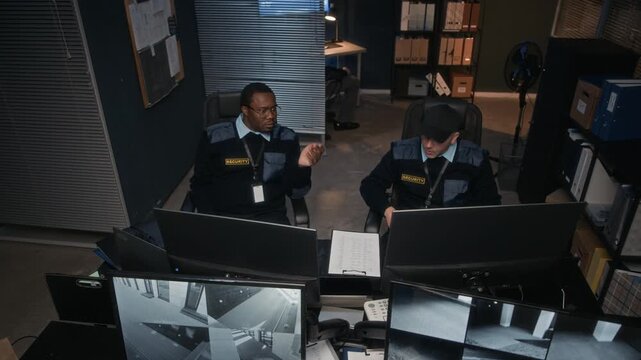 Multiethnic team of three security guards reacting quickly to red alert on computer screens during night shift in control room, one communicating with walkie talkie