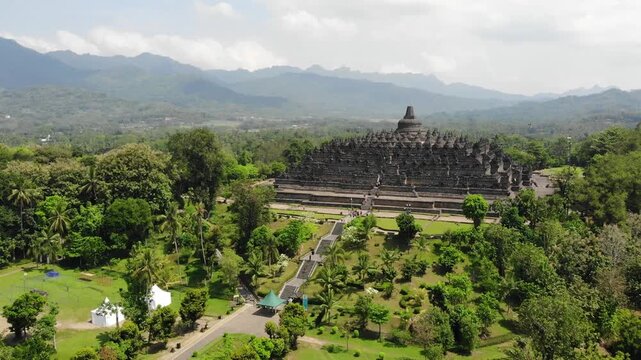 Aerial Tilt Up Wide Shot of Borobudur Temple with Scenic Mountain Background Under Partly Cloudy Sky in 4K. Cinematic wide aerial drone view of the Borobudur Temple in Indonesia.